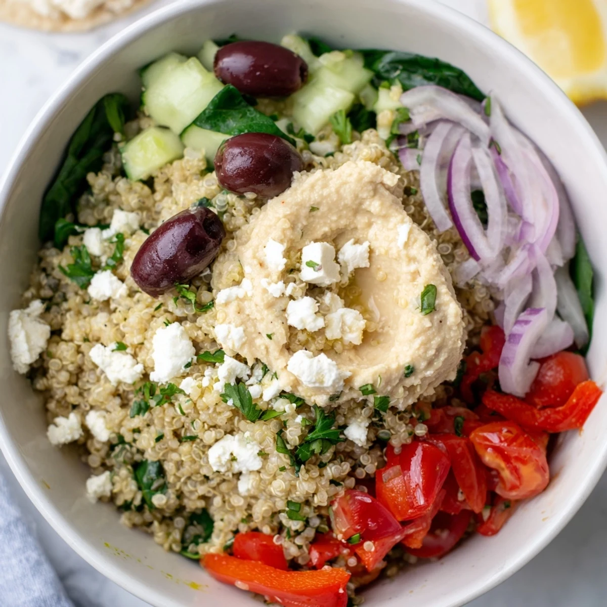 Vibrant Mediterranean Quinoa Bowl topped with creamy hummus, briny olives, and crumbled feta beside fresh cherry tomatoes and cucumber.