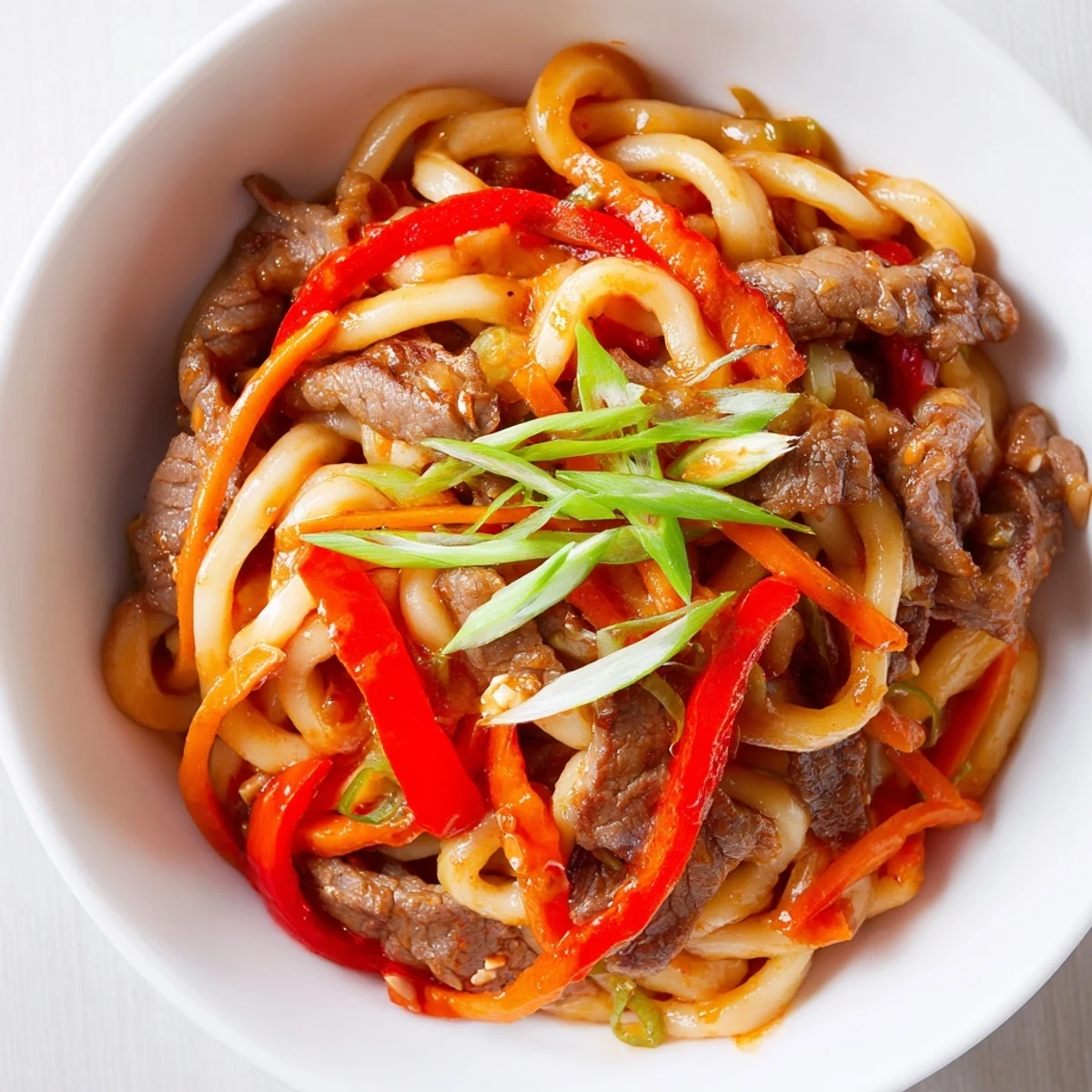 A plated serving of Spicy Beef Stir Fry with Udon Noodles, garnished with spring onions and a drizzle of sauce.