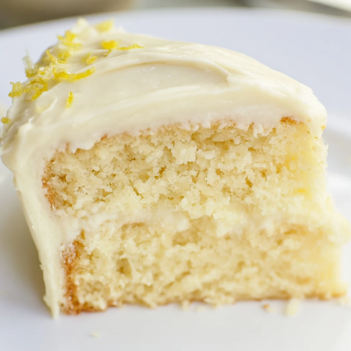 A moist lemon cake slice with luscious lemon cream cheese frosting, topped with zest curls and served on a white plate.