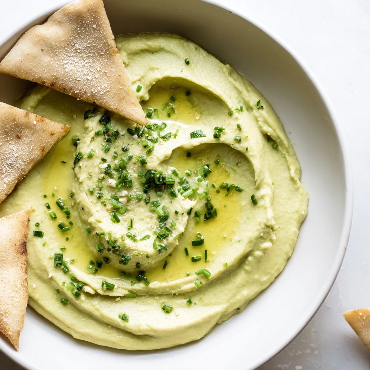 A bowl of vibrant green avocado hummus topped with olive oil, surrounded by golden, crunchy homemade pita chips.  