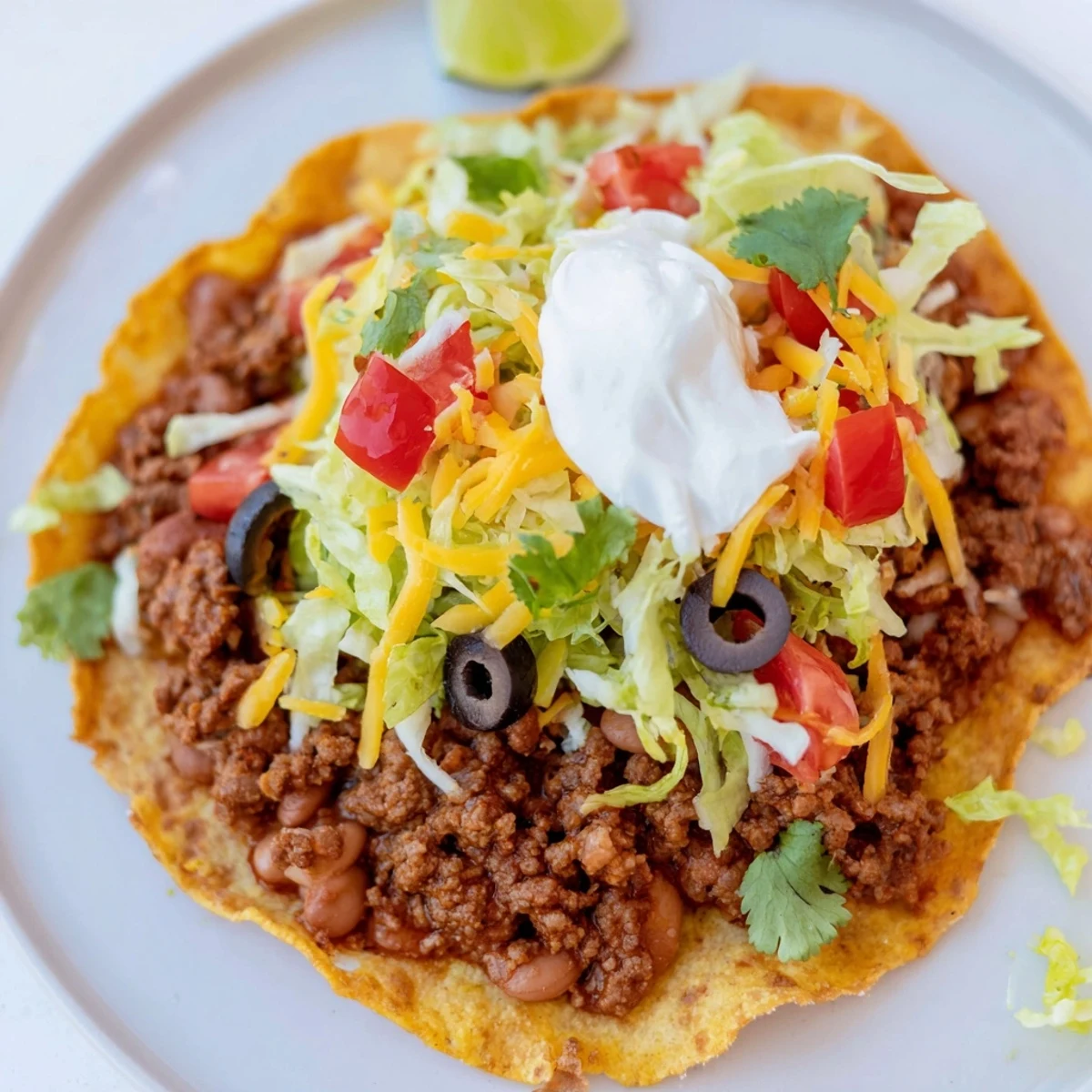 A close-up view of Beef Tostadas with Refried Beans showcasing melted cheese, sliced black olives, and a dollop of sour cream on top.