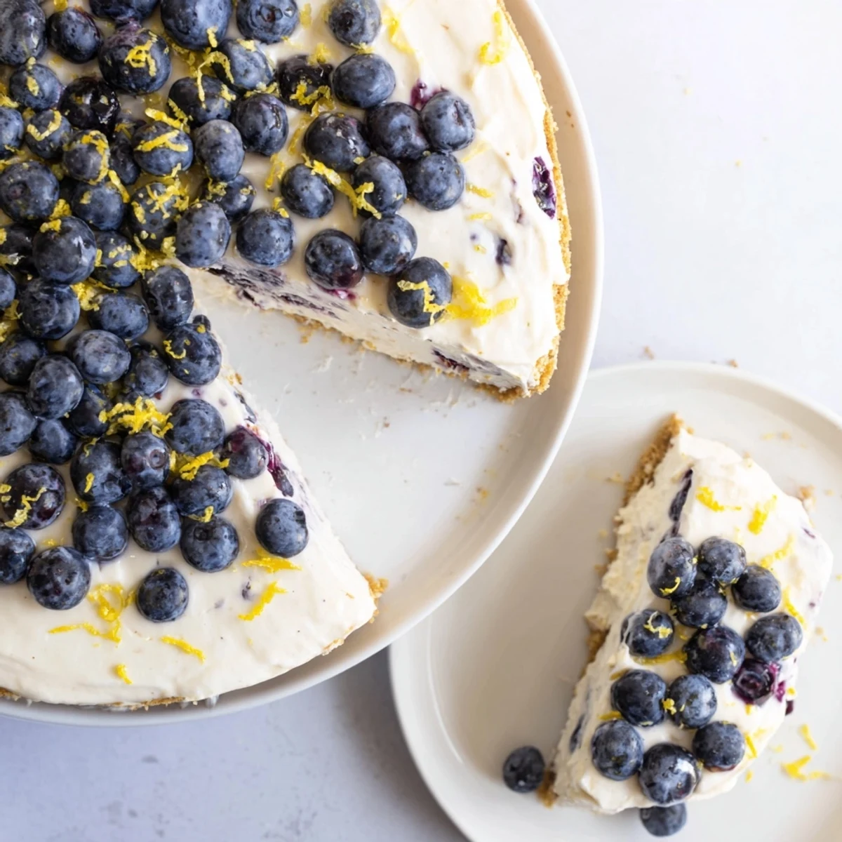 A chilled no bake lemon blueberry cream cake reveals graham cracker crumbs, lemon zest, and blueberries on a white plate.